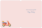 Inside of a light pink birthday card, showing the pre-printed message 'Here's to living the good life! Happy Birthday' in blue script, with a small scooter illustration in the bottom left corner.