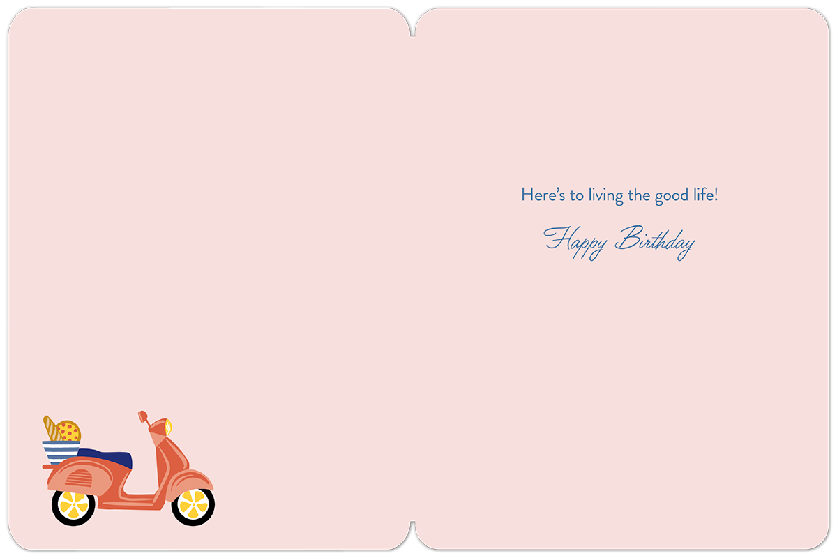 Inside of a light pink birthday card, showing the pre-printed message 'Here's to living the good life! Happy Birthday' in blue script, with a small scooter illustration in the bottom left corner.