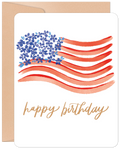 Front of a patriotic greeting card featuring a watercolor American flag with blue floral stars and red wavy stripes, accompanied by the text 'happy birthday' in gold script, resting on a brown kraft envelope.