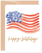 Front of a patriotic greeting card featuring a watercolor American flag with blue floral stars and red wavy stripes, accompanied by the text 'happy birthday' in gold script, resting on a brown kraft envelope.