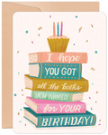 Greeting card featuring a stack of colorful books topped with a cupcake and lit candles, with text 'I hope YOU GOT all the books YOU WANTED for YOUR BIRTHDAY!' on a light background with confetti.