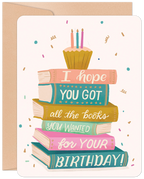 Greeting card featuring a stack of colorful books topped with a cupcake and lit candles, with text 'I hope YOU GOT all the books YOU WANTED for YOUR BIRTHDAY!' on a light background with confetti.