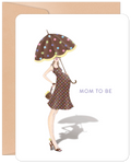 Front of a cute 'Mom to Be' greeting card featuring an illustration of a stylish pregnant woman in a polka dot dress and holding a matching umbrella, on a white background with a kraft envelope.