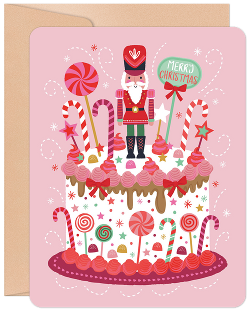Pink Christmas greeting card featuring a whimsical illustration of a nutcracker soldier standing on a large, decorated cake with candy canes, lollipops, frosting, and sprinkles, surrounded by snowflakes and a 'Merry Christmas' speech bubble.
