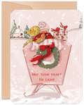 Adorable golden retriever puppy in a winter hat riding a vintage pink sled with festive gifts, a holiday wreath, and falling snow. Front of a heartfelt greeting card featuring a cozy winter village background and the text 'MAY YOUR HEART BE LIGHT'.
