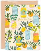 A square greeting card featuring a vibrant pattern of lemons, oranges, and greenery in mason jars and soda cans, with the gold script text "You're the Zest!". A kraft paper envelope is partially visible behind the card.
